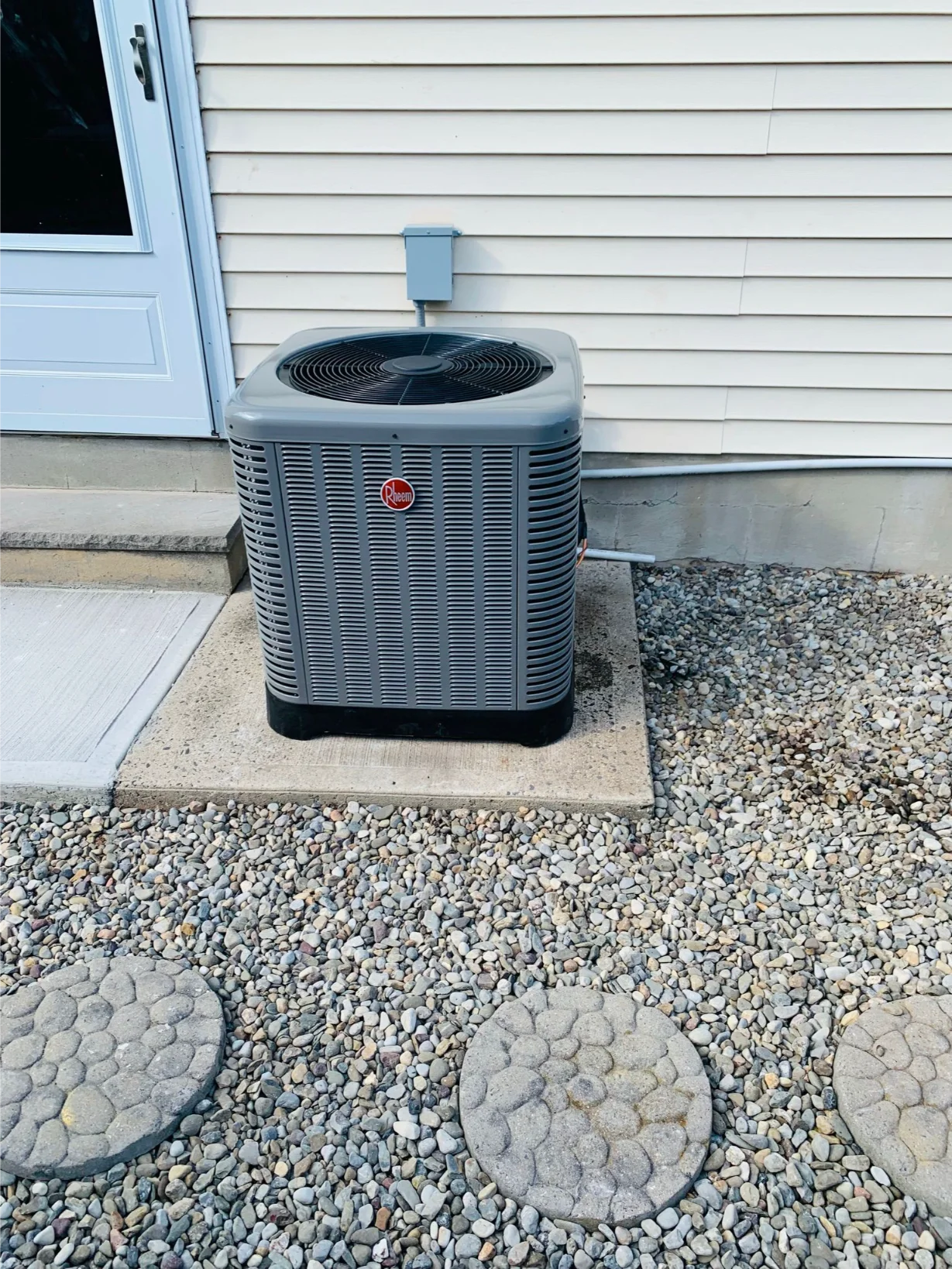 newly installed Rheem outdoor AC unit next to a house entrance, with decorative stone path
